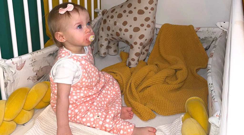 Kislány cumival rózsaszín tipegő hálózsákban ül a kiságyában