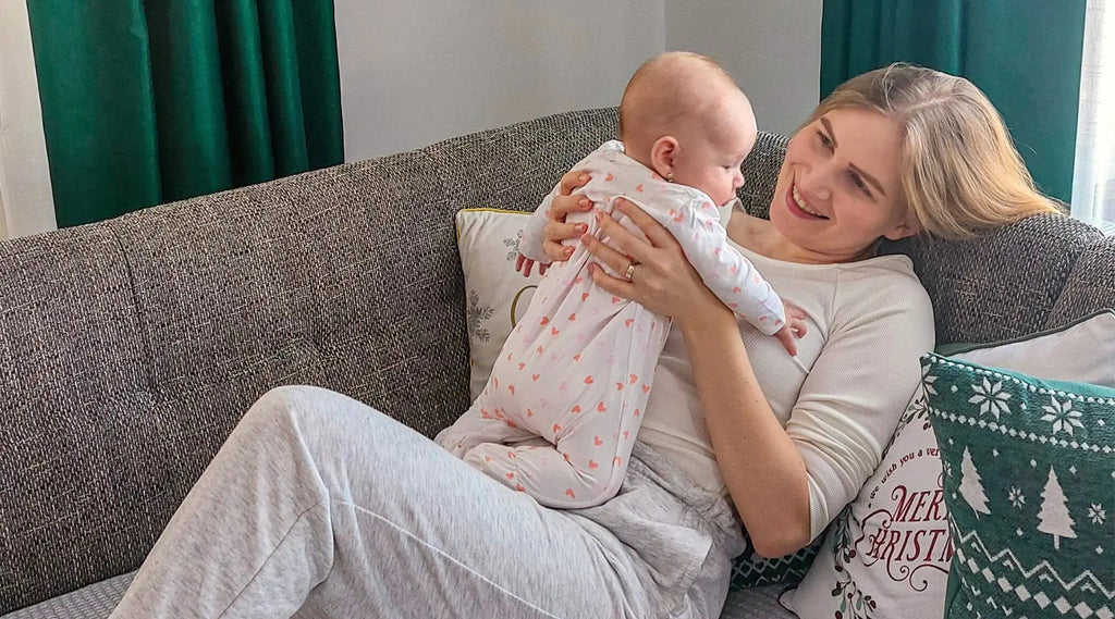 Anya fogja a kisbabáját a kezében a kanapén fekve és rá mosolyog.