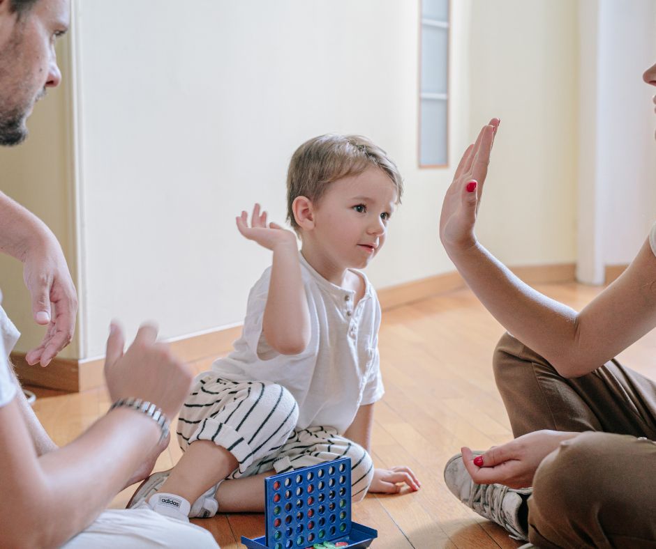 Apa, anya és kisfiuk a padlón játszanak, a gyermek éppen pacsizik az anyukájával, ami a harmonikus családi kapcsolatot és a pozitív nevelést jelképezi.